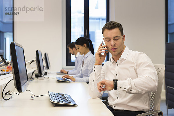 Mann am Schreibtisch am Handy mit Kollegen im Hintergrund