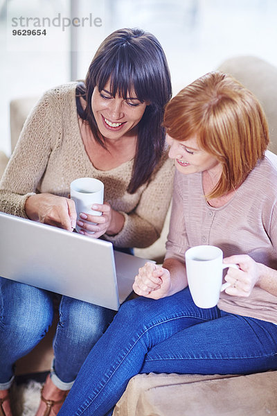 Zwei glückliche Frauen sitzen auf der Couch mit Laptop und Tassen