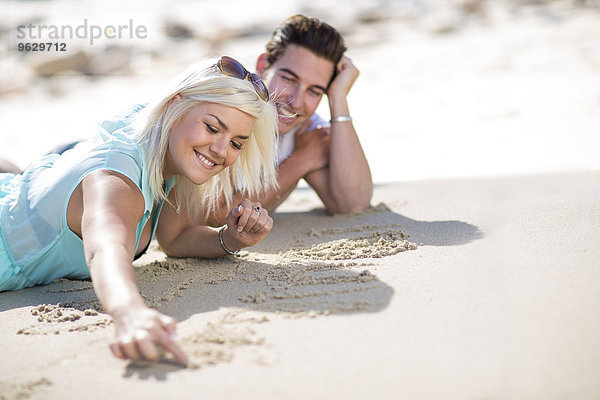 Junges Paar am Strand liegend und im Sand zeichnend