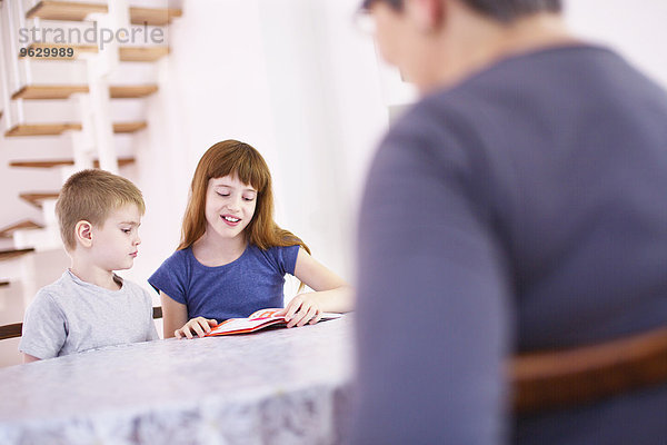 Großmutter beobachtet Enkelkinder beim Lesen am Küchentisch