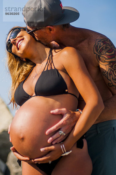 Reifer Mann  der seine schwangere Frau am Strand umarmt und küsst.