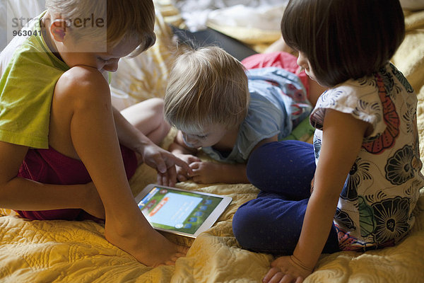 Drei kleine Kinder entspannen sich auf dem Bett mit Hilfe eines digitalen Tabletts.