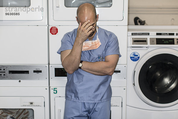 Älterer Mann in chirurgischen Peelings mit Hand auf der Stirn in der Waschküche