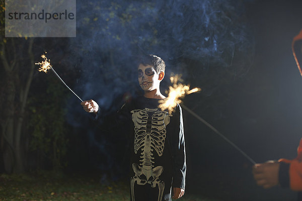 Junge im Halloween-Skelettkostüm mit Wunderkerze im Garten bei Nacht