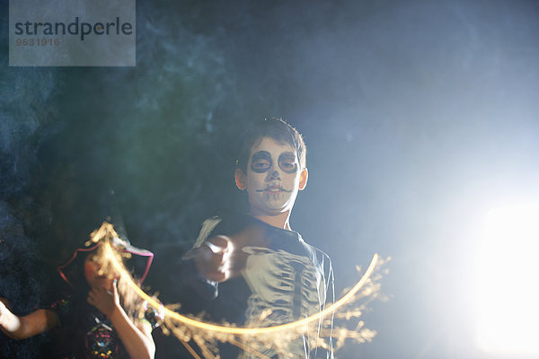 Geschwister in Halloween-Kostümen spielen nachts mit Wunderkerzen im Garten.