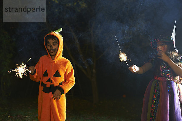 Geschwister in Halloween-Kostümen mit Wunderkerzen im Garten bei Nacht