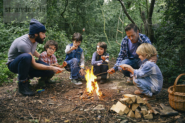 Zwei Väter und vier Jungen stoßen auf Marshmallows am Lagerfeuer im Wald an.