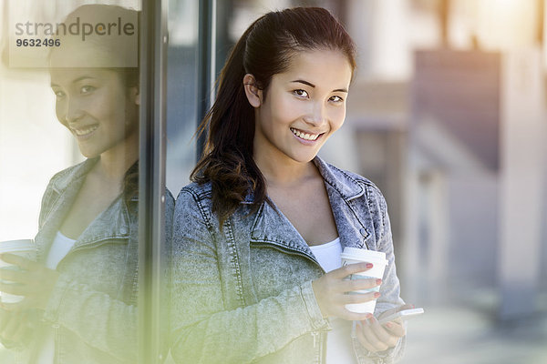 Porträt einer jungen Frau mit Kaffee zum Mitnehmen und Smartphone  das sich gegen das Parkhaus lehnt