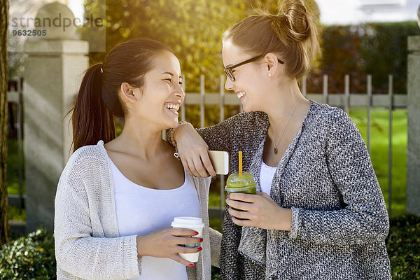 Zwei junge Freundinnen im Park  die lachen und Kaffee zum Mitnehmen trinken.