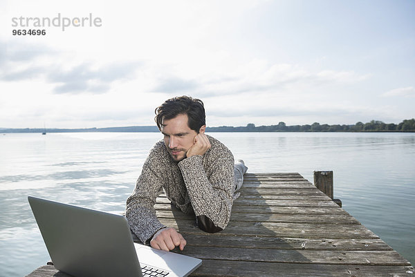Young man lying on wooden jetty lake laptop