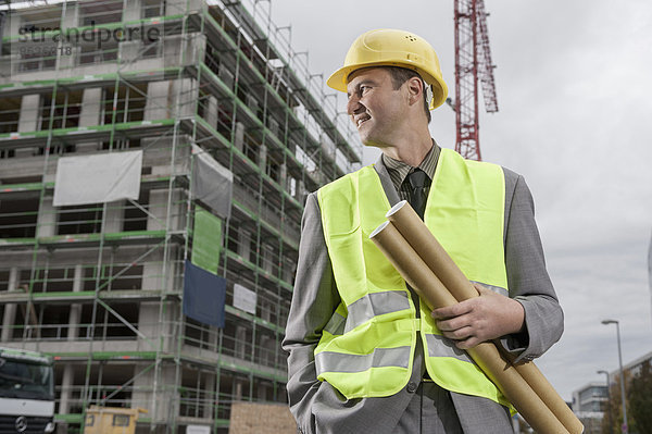 Happy male building contractor standing with blueprints and hands in pockets at a construction site