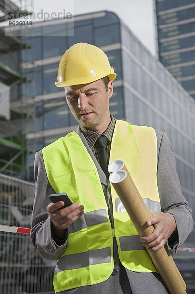 Site manager holding blueprints and text messaging on a mobile phone