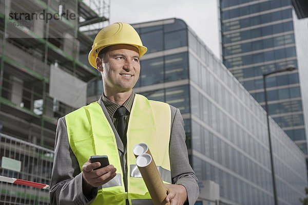 Happy site manager holding blueprints and mobile standing at a construction site