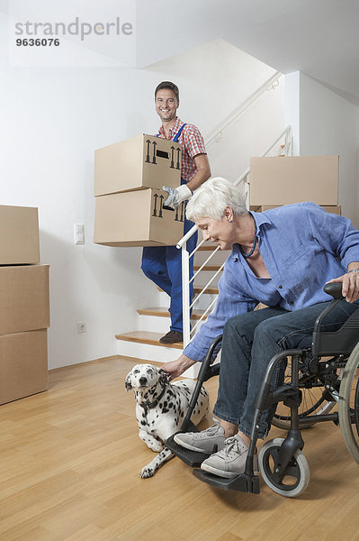 Handicapped old woman stroking dog while movers walking down cardboard boxes