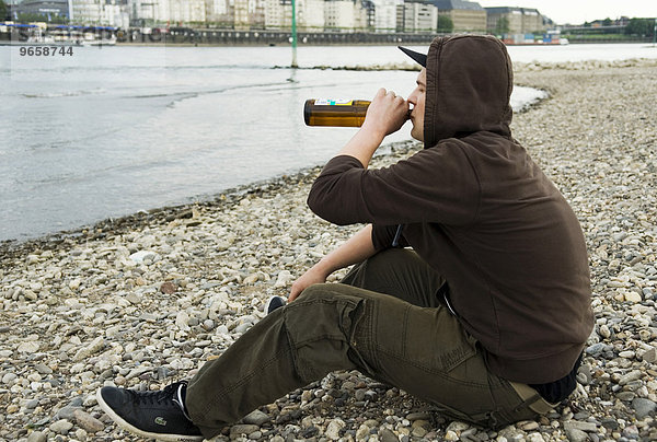 Einsamer Jugendlicher sitzt am Rheinufer und trinkt eine Flasche Bier