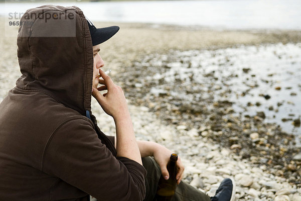 Einsamer Jugendlicher sitzt am Rheinufer  raucht und trinkt eine Flasche Bier