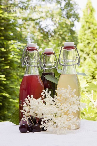 Drei Glasflaschen selbstgemachter Sirup auf Gartentisch: Rhabarbersirup  Kirschsirup  Holunderblütensirup