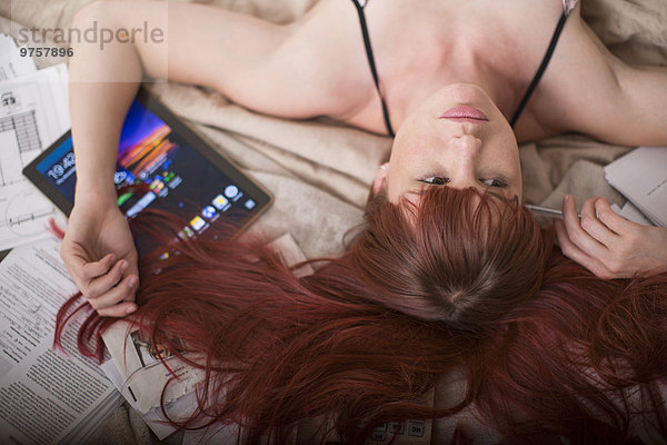 Frau auf dem Bett liegend mit digitalem Tablett und Papierkram