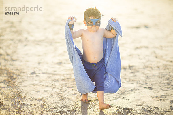 Kleiner Junge am Strand  verkleidet als Superheld mit Maske und Handtuch.