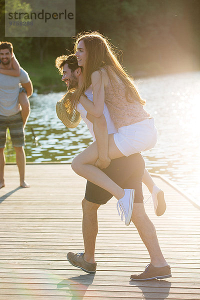 Fröhlicher junger Mann trägt Freundin Huckepack am Steg am See
