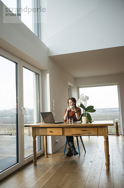 Junge Frau mit Laptop am Holztisch mit Blick aus dem Fenster