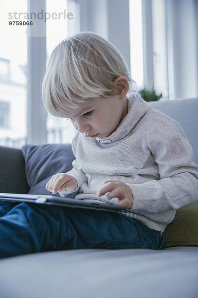 Kleiner Junge sitzt auf der Couch und spielt mit dem digitalen Tablett.