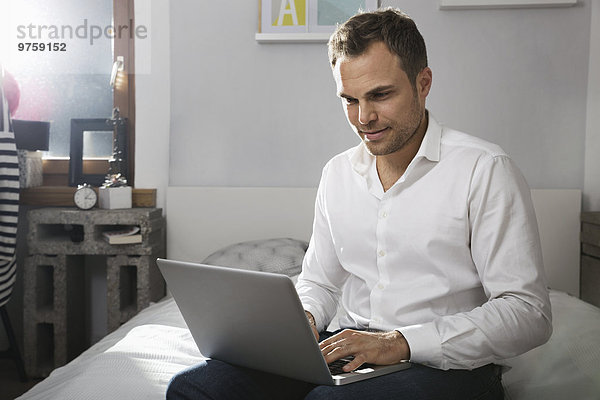 Mann sitzt auf dem Bett zu Hause mit Laptop