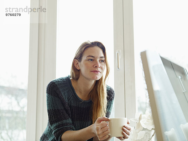 Deutschland  Köln  Junge Frau am Fenster sitzend  Tasse Kaffee haltend