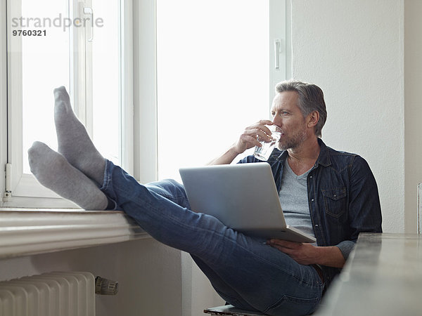 Deutschland  Köln  reifer Mann am Fenster sitzend mit Laptop  Füße oben