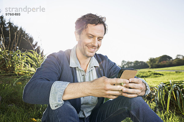 Lächelnder Mann sitzt auf einer Wiese und liest SMS
