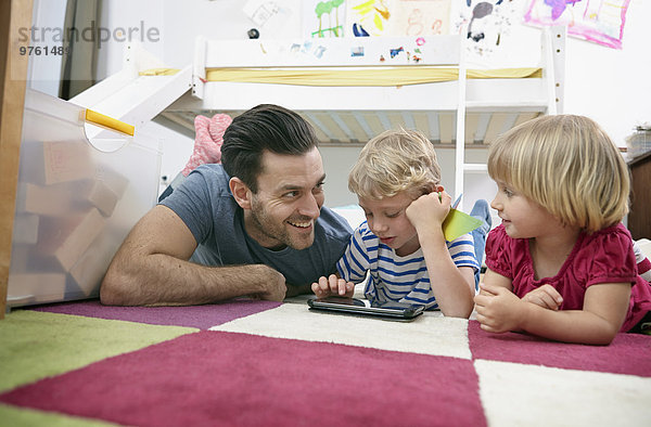 Vater und Kinder mit Mini-Tablette  auf dem Boden liegend
