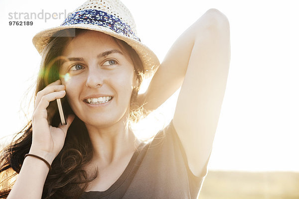 Lächelnde Frau mit Sommerhut telefoniert mit Smartphone