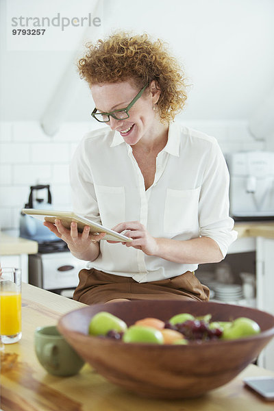 Porträt einer Frau  die in der Küche sitzt und auf das digitale Tablett schaut  lächelnd
