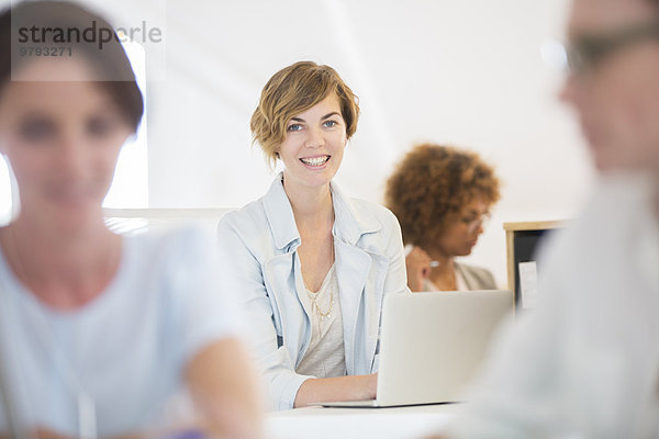 Porträt einer Frau am Schreibtisch sitzend  mit Laptop und lächelnd im Büro  Kollegen im Hintergrund