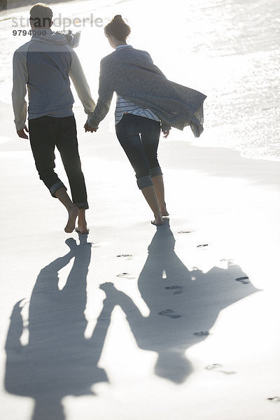 Junges Paar hält sich an den Händen und geht am Strand spazieren.