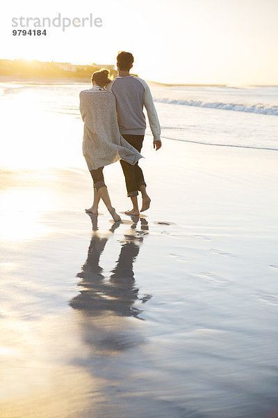 Junges Paar hält sich an den Händen und geht bei Sonnenuntergang am Strand spazieren.