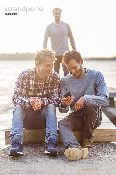 Glückliche männliche Freunde mit Handy am Pier mit Mann im Hintergrund