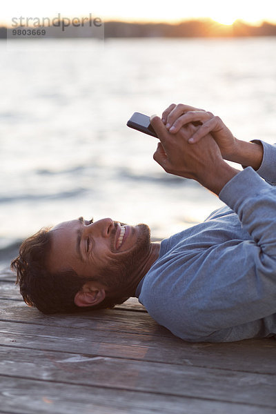 Glücklicher Mann  der bei Sonnenuntergang auf einem hölzernen Pier gegen das Meer liegt und ein Handy benutzt.