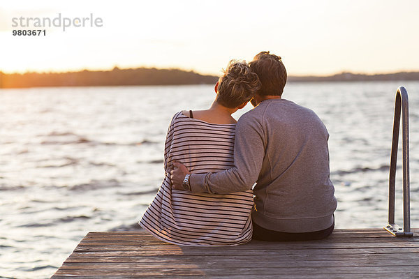 Rückansicht des liebevollen Paares  das bei Sonnenuntergang auf dem Pier gegen das Meer sitzt.