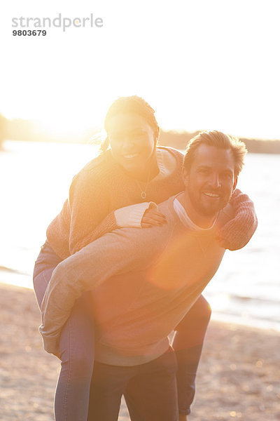 Porträt eines liebevollen Paares  das bei Sonnenuntergang Huckepackfahrt am Strand genießt.
