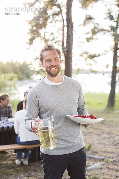 Porträt eines glücklichen Mannes mit Bierkrug und Erdbeeren mit Freunden am Picknicktisch im Hintergrund