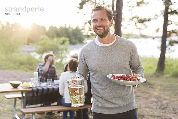 Glücklicher Mann mit Bierkrug und Erdbeeren mit Freunden am Picknicktisch im Hintergrund