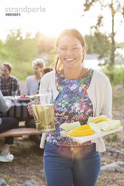 Porträt einer glücklichen Frau mit Bierkrug und Zuckermelonenscheiben mit Freunden am Picknicktisch im Hintergrund.