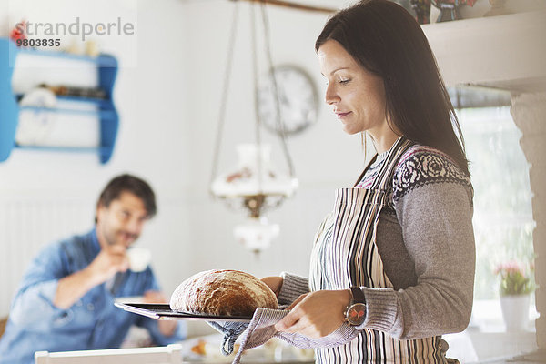 Frau mit frisch gebackenem Brot im Tablett und Mann im Hintergrund zu Hause