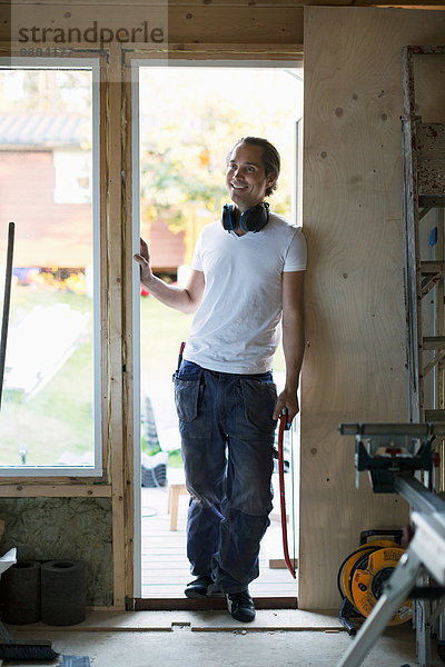 Volle Länge des lächelnden Mannes  der sich in der Tür des zu renovierenden Hauses lehnt.