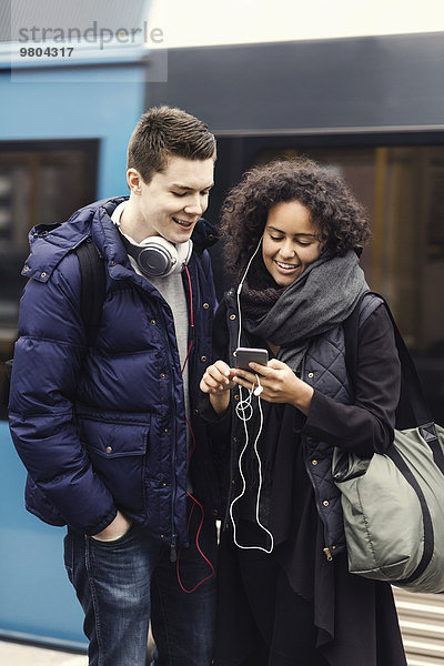 Multi-ethnisches Paar  das Musik über Handy auf der U-Bahn-Plattform hört.