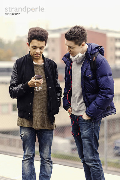 Männliche Universitätsstudenten beim Warten auf dem U-Bahn Bahnsteig mit dem Handy