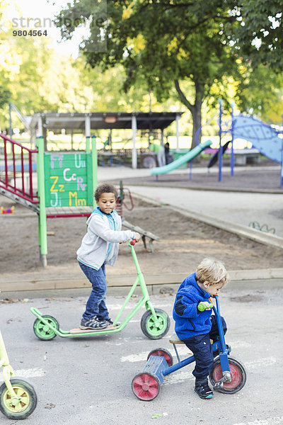 Junge mit Roller  der auf dem Spielplatz einen Freund beim Dreiradfahren ansieht.