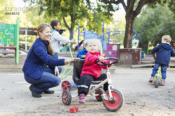 Glückliche Lehrerin  die dem Mädchen hilft  auf dem Spielplatz Dreirad zu fahren.