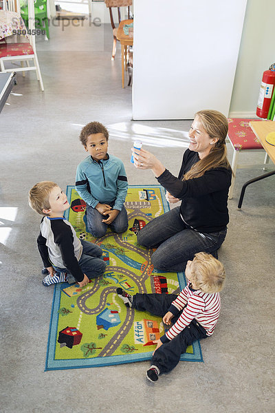 Hochwinkelansicht der glücklichen Lehrerin mit Blasenlösung beim Sitzen mit Kindern im Kindergarten